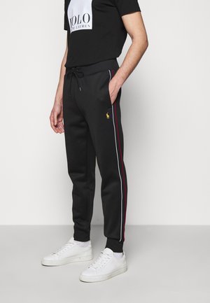 Mand iført sorte træningsbukser med røde og hvide sidelstriber, gul logo på låret, sort snøre ved taljen, hvide sneakers og sort Polo Ralph Lauren t-shirt.