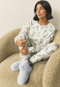 Valkoinen pyjama, jossa mustia viivapiirroksia, pitkät hihat ja joustavat kalvosimet. Pehmeät harmaat pörrösiset tohvelit. Istuessaan teksturoidulla beigenvärisellä tuolilla.