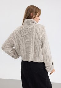 Pikakarvade ja mustade kõrgete vöödega pükstega naine, kellel on pikad juuksed ja seljas beež jäme kaelus-sweater, millel on punutud kudumise mustrid, pöörab külje poole.