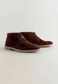 Stivali alla caviglia in suede marrone con lacci abbinati, punta tonda e suola leggera con dettagli di cucitura lungo i bordi.