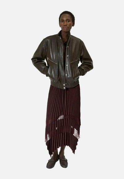 Mujer con chaqueta de cuero marrón oscuro, falda burgundy plisada y asimétrica, y mocasines marrones a juego, de pie con las manos en los bolsillos.