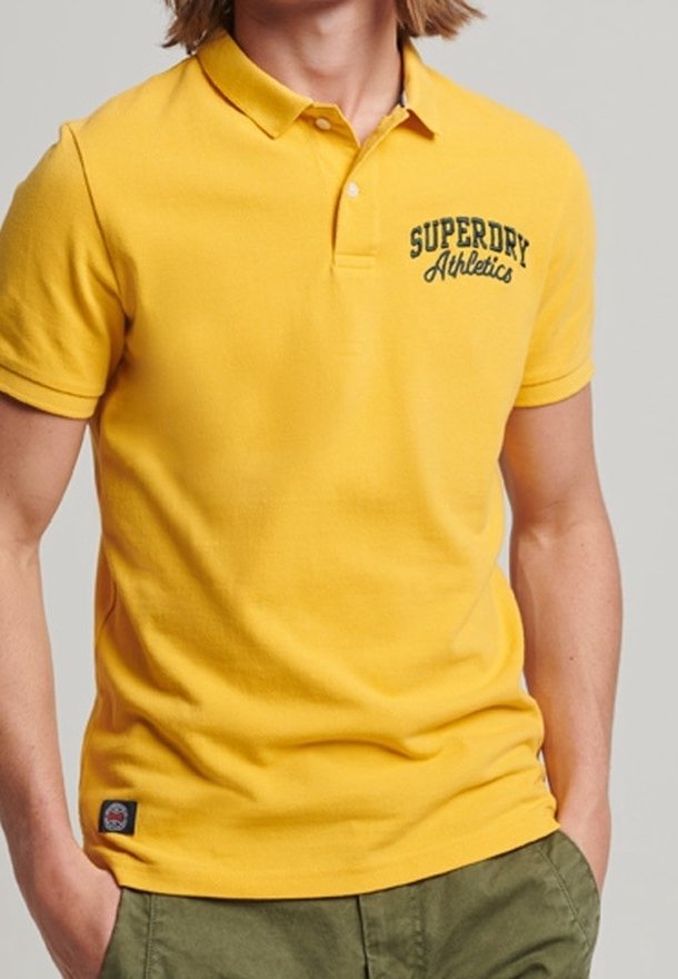 Homme portant un polo jaune Superdry Athletics avec col et boutons, associé à un pantalon vert olive, les mains dans les poches.