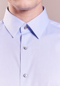 Světle modrá košile na knoflíky má strukturovaný límec, texturovanou tkaninu a stříbrné knoflíky s hladkým povrchem.