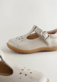 Beżowe lakierowane buty na obcasie z paskiem T, zapięciem na klamrę i ozdobnym perforowanym detalem na palcach. Gumowa podeszwa z teksturowanym wzorem.