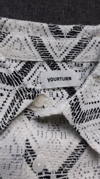 Camicia in pizzo con un motivo geometrico bianco e nero, dotata di chiusura con bottoni e un'etichetta con la scritta "YOURTURN." Tessuto dettagliato e testurizzato.