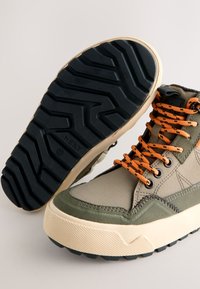 Chaussures montantes vertes et beiges avec une semelle en caoutchouc texturée, des lacets orange et un mélange de toile et de tissus synthétiques.