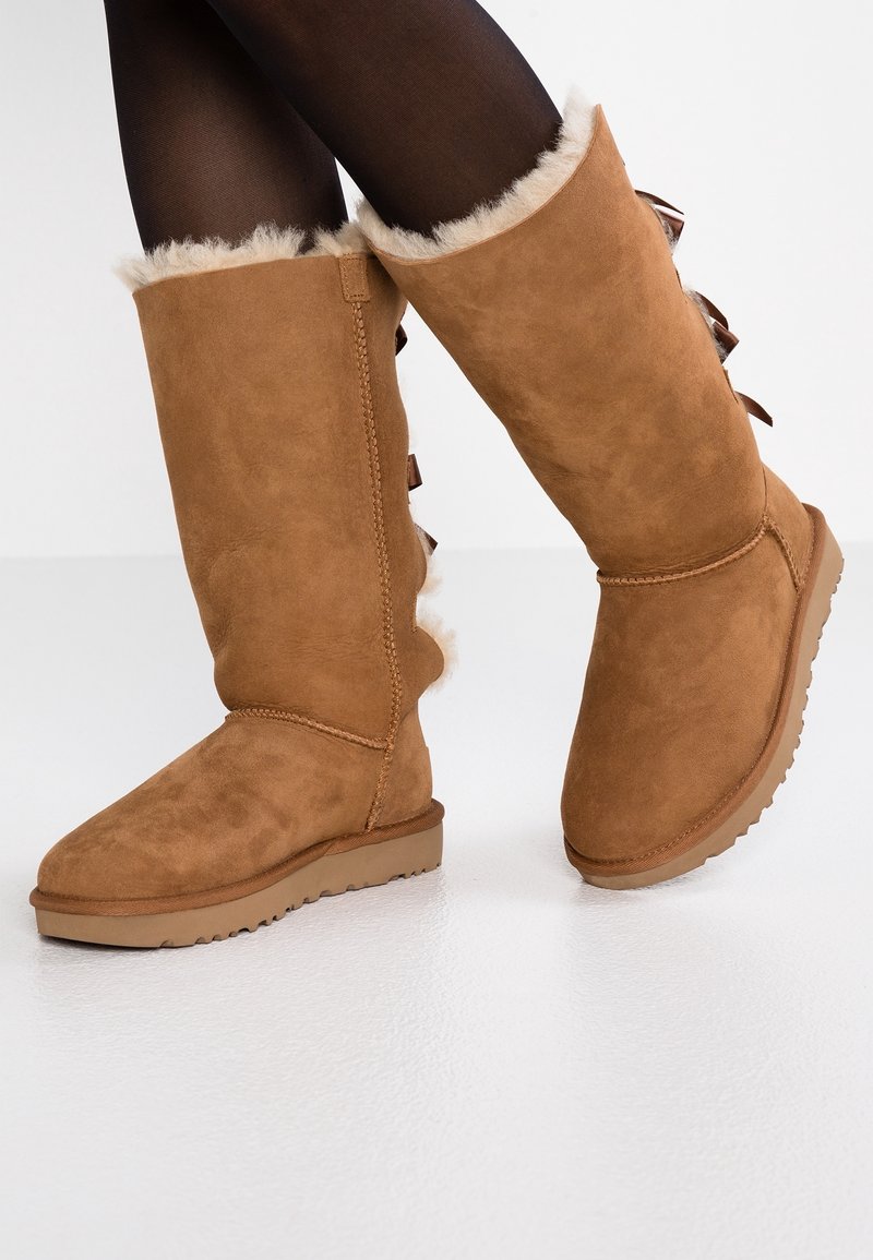 Bottes montantes en suède marron avec bordure en fausse fourrure en haut, dotées d'une semelle en caoutchouc texturée et de nœuds décoratifs à l'arrière.