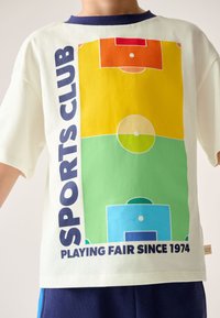 Balti marškinėliai su spalvotu futbolo aikštės dizainu, kuriame yra žali, geltoni, oranžiniai ir mėlyni plotai; tamsiai mėlynas apykaklė.