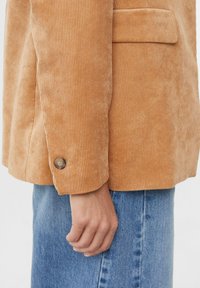 Sztruksowa marynarka w jasnobrązowym kolorze o fakturowanym wykończeniu. Wyposażona w mankiety zapinane na guziki oraz kieszeń na piersi. Połączona z dżinsową spódnicą.