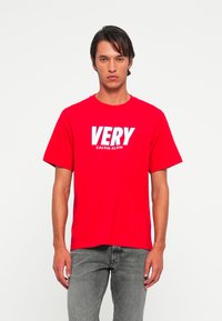 Vyras, stovintis prieš baltą foną, dėvi raudoną marškinėlį su užrašu "VERY CALVIN KLEIN" ir pilkas džinsus.