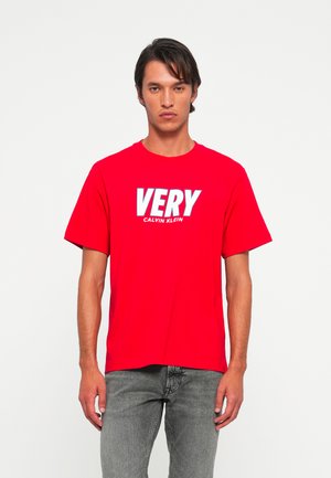 Vyras, stovintis prieš baltą foną, dėvi raudoną marškinėlį su užrašu "VERY CALVIN KLEIN" ir pilkas džinsus.