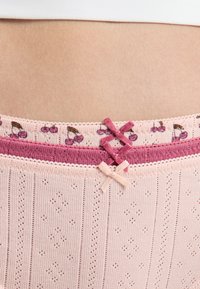 Różowa koronka z wzorem wiśni, dwa małe kokardki w ciemniejszych odcieniach różu na pasku. Tkanina teksturowana z małymi otworami i liniami.