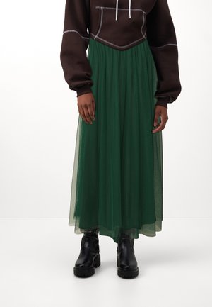 Person iført en mørkebrun cropped hættetrøje, lang grøn tylskørt og sorte chunky ankelstøvler, stående mod en hvid baggrund.