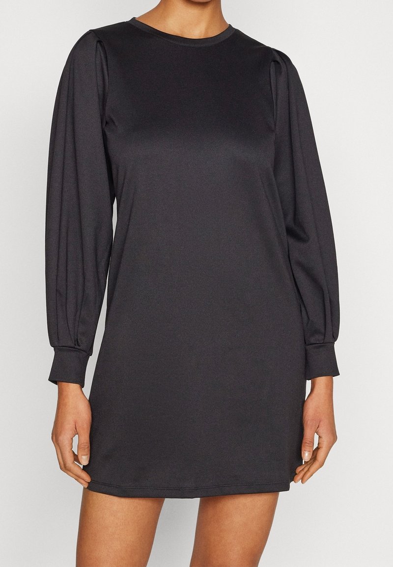 Femme portant une mini robe noire à manches longues légèrement bouffantes avec des poignets ajustés, se tenant devant un fond uni.