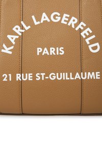 Tote bag i lyst tan-skinn med hvit preget tekst: "KARL LAGERFELD," "PARIS," og "21 RUE ST-GUILLAUME." Teksten plassert i en bue.
