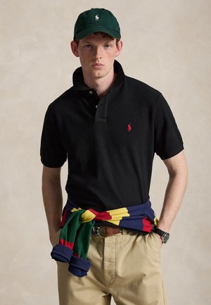 Ung mand iført en mørkegrøn baseballkasket, sort poloshirt med rød logo, beige bukser, ur og en flerfarvet sweater bundet om taljen.