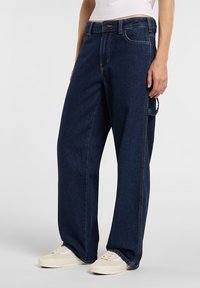 Dunkelblaue, weite Jeans aus Denim mit hohem Bund, versehen mit Taschen vorne und hinten sowie gelben Nähten. Kombiniert mit weißen Turnschuhen.