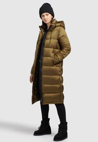 Long manteau doudoune vert olive avec capuche, fermeture éclair sur le devant, deux poches latérales et un design matelassé. Porté avec des leggins noirs et un bonnet tricoté noir ainsi que des bottes.