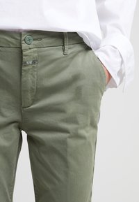 Närbild av person i olivgröna byxor med framknapp och hand i fickan, iklädd en vit långärmad skjorta med manschett.