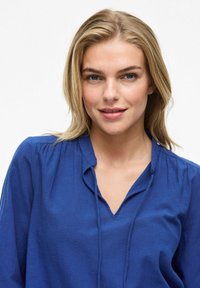 Maglietta blu a scollo a v con maniche lunghe, realizzata in un materiale morbido. Presenta una vestibilità rilassata e pieghe sottili sulle spalle.