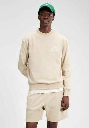 Young man wearing beige sweatshirt and shorts with white undershirt, green cap, standing against light gray background.