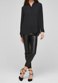 Zwarte langärmige blouse met knopen en een zachte textuur, gecombineerd met strakke zwarte leren broeken en spitse zwarte hakken met enkelbandjes.