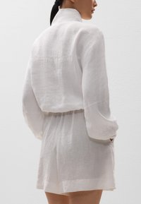 Weißes Leinenoutfit mit hochgeschlossenem Kragen, langen Ärmeln und lockerer Passform. Hat eine taillierte Taille und kurze, locker sitzende Shorts.