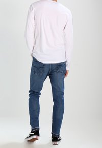 Persona de espaldas, vistiendo una camisa blanca de manga larga, pantalones vaqueros azules y zapatillas negras bajas con suelas blancas.