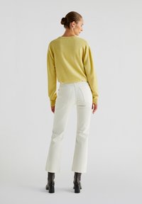 Geel gebreide trui met lange mouwen, gecombineerd met witte uitlopende jeans. Model draagt kniehoge zwarte haklaarzen. Eenvoudig, minimalistisch ontwerp.