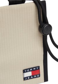 Γωνία μπεζ υφασμάτινης τσάντας με μαύρο περιθώριο, μαύρο κορδόνι σύσφιξης και ετικέτα λογότυπου Tommy Jeans σε κόκκινα, λευκά και ναυτικά χρώματα.