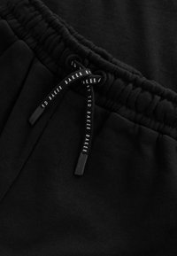 Sorte joggingbukser med snørebånd i taljen. Snørebåndet har trykt tekst, og stoffet har en glat tekstur.