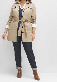 Beige trenssitakki, jossa vyö, napillinen suljenta ja olkatoppa, kerroksittain päällä tummansininen neuletakki ja valkoinen paita, yhdistetty tummiin farkkuihin ja ruskeisiin saappaisiin.