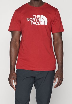 Mand iført en rød T-shirt med korte ærmer med stort hvidt "The North Face" logo og sorte bukser, stående mod en ensartet baggrund.