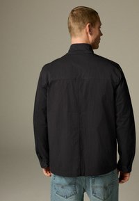 Uomo che indossa una giacca nera a maniche lunghe e jeans azzurri chiari, di spalle contro uno sfondo neutro e semplice.