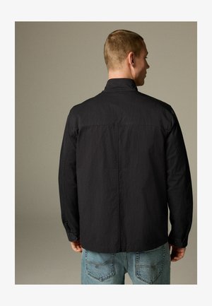 Férfi fekete hosszúujjú dzsekiben és világoskék farmernadrágban, háttal áll egy sima semleges háttér előtt.