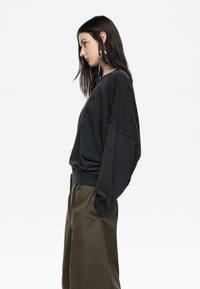 Čierny oversized sweatshirt s klesnutými ramenami, textúrovaná tkanina a nenápadné logo. V kompletne s olivovozelenými nohavicami širokého strihu.