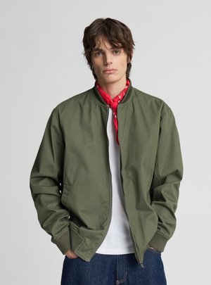 Młoda osoba w oliwkowozielonej kurtce na zamek, białej koszuli, czerwonym chusteczce i niebieskich dżinsach, stojąca z rękami w kieszeniach.