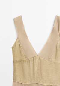 Beige hihaton mekko, jossa on leveät olkaimet ja syvä V-aukkoinen kaula-aukko, jossa on tekstuuroitu kangaspaneeli rinnassa.
