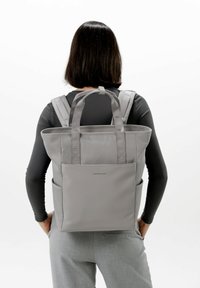 Grå tote-stil ryggsäck med dubbla remmar, texturerad tyg och två sidofickor. Har en minimal logotyp på frontpanelen.