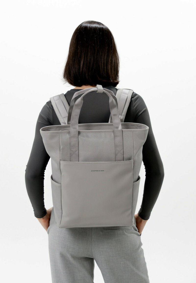 Grå tote-stil ryggsäck med dubbla remmar, texturerad tyg och två sidofickor. Har en minimal logotyp på frontpanelen.