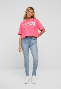 Rózsaszín, cropped póló merész fehér felirattal, világoskék skinny farmernadrággal és fehér tornacipővel párosítva. Egyszerű, hétköznapi öltözet sima háttér előtt.