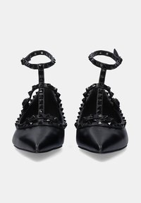 Zapatos negros de punta afilada con acentos de tachuelas, correas alrededor del tobillo y una textura de cuero suave. Presentan herrajes decorativos a lo largo de los bordes.