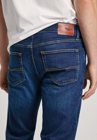 Jeans de mezclilla azul oscuro con una textura suave. Las características incluyen dos bolsillos traseros con costuras amarillas y una etiqueta de cuero marrón en la cintura.