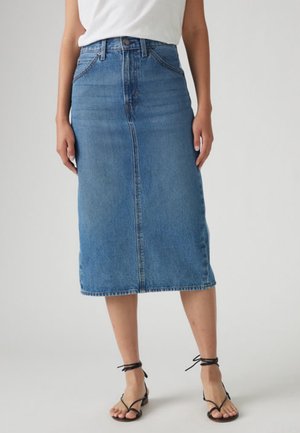 Femme portant une jupe en jean bleue longueur genou, un haut blanc et des sandales à talons noires à lanières sur un fond uni.