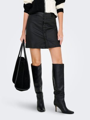 Femme portant une mini-jupe en cuir noire boutonnée à l'avant, des bottes noires à talons montant jusqu'aux genoux, tenant un grand sac cabas noir dans la main gauche.