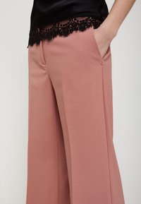 Brede pijpenbroek van zacht roze stof, met een zwarte kantafwerking bij de taille. Inclusief zijzakken en subtiele knoopdetails.