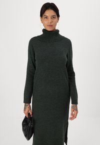 Abito lavorato a maglia verde scuro con colletto alto a coste, maniche lunghe, spacchi laterali e una consistenza morbida. Il modello tiene una pochette nera.