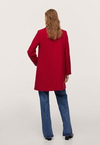 Femme aux longs cheveux portant un manteau rouge, un jean bleu et des chaussures à talons noires, se tenant de dos contre un fond uni.