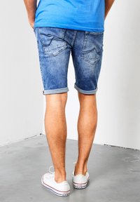 Pantalones cortos de mezclilla en azul claro con un acabado deslavado, puños doblados y dos bolsillos traseros con detalles de bordado curvados. Zapatillas blancas.