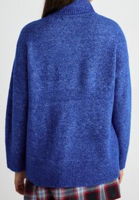 Maglione blu oversize con collo alto, lavorazione a maglia testurizzata, polsini e orlo a costine. Indossato sopra una gonna scozzese con più colori.